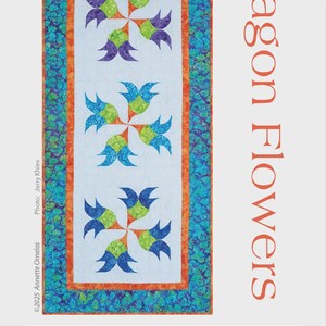 May include: A rectangular quilt with a floral design. The quilt features a white center with colorful flower patterns in blue, green, and purple. The border is a mix of orange and teal. The text "Dragon Flowers" is displayed vertically. The table runner is 24" x 54".