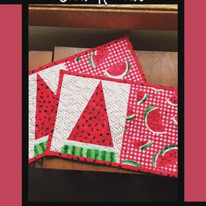 May include: Two watermelon-themed snack mats. The mats feature a red and white color scheme with a watermelon slice design. The text "Just One Slice Snack Mat" is at the top. The mats are on a wooden surface.