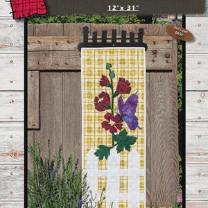 May include: A quilted wall hanging with the text "HOLLYHOCK HAVEN" and dimensions of 12" x 31". The design features a yellow plaid background, a white picket fence, and a floral and butterfly motif in shades of purple, red, and green.