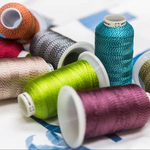 May include: A collection of spools of colorful embroidery thread, including green, purple, brown, gray, blue, and orange. The spools are arranged on a white surface.