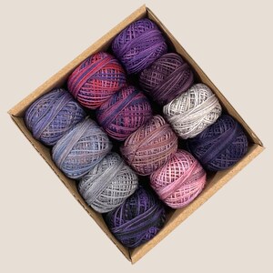 May include: A cardboard box filled with twelve yarn balls in various shades of purple, pink, and gray. The yarn balls are tightly wound and arranged in a grid pattern. The box is angled, showing the different colors and textures of the yarn.