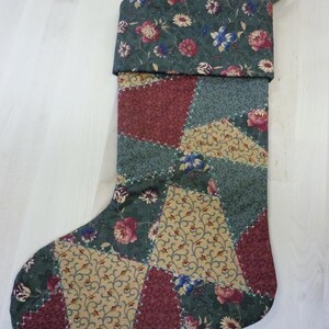 Op de afbeelding: Een gewatteerde kerstsok met een patchworkontwerp van driehoekige stofsegmenten in tinten donkergroen, bordeauxrood en beige. De stof toont bloemmotieven en gedetailleerd krulwerk, met een bijpassende bloemrijke boord en een stoffen ophanglus.