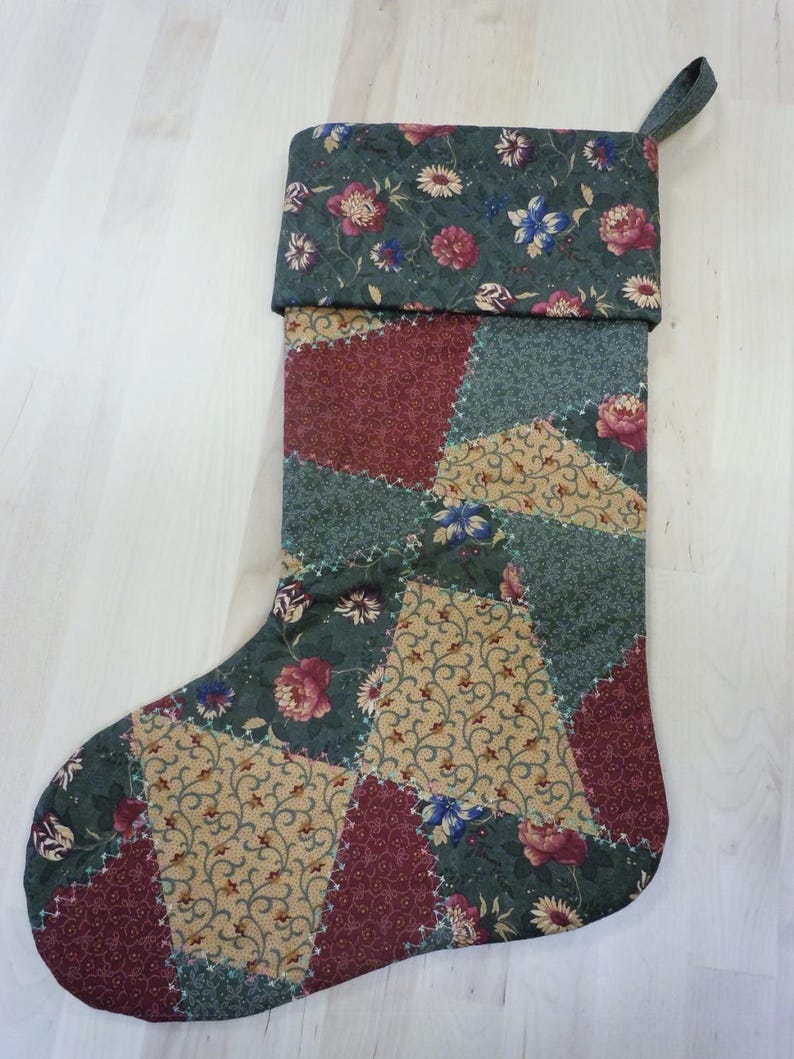 Op de afbeelding: Een gewatteerde kerstsok met een patchworkontwerp van driehoekige stofsegmenten in tinten donkergroen, bordeauxrood en beige. De stof toont bloemmotieven en gedetailleerd krulwerk, met een bijpassende bloemrijke boord en een stoffen ophanglus.
