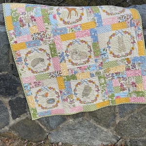 May include: A patchwork quilt featuring embroidered woodland animals within floral wreaths. The quilt has a variety of pastel-colored squares and the text "Backyard Buddies". The quilt is made by Sweet Jane's Quilting & Design.