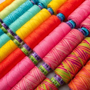 May include: A close-up of a variety of colorful embroidery floss spools. The spools are arranged in a row, with the colors transitioning from green to pink to orange. The spools are labeled with the brand name "WonderFil" and the thread number.