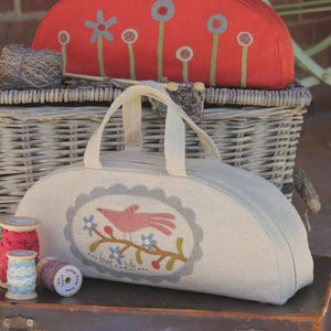 May include: Two half-circle shaped fabric bags with handles. The top bag is red with a flower and stem design. The bottom bag is beige with a bird and branch design. The text "love...life..." is embroidered on the bag. The bags are sitting on top of a brown suitcase.