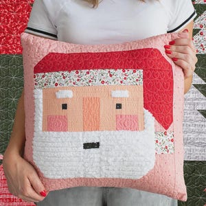 May include: A decorative Christmas pillow featuring a Santa Claus face. The pillow has a pink background with a red hat and beard. The Santa face is made of various fabrics, including white, pink, and red. The pillow is quilted.