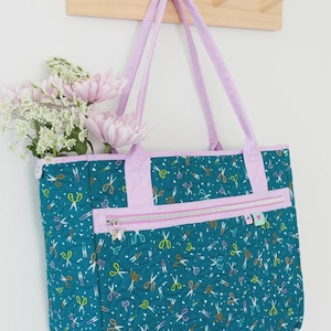 May include: A teal quilted tote bag featuring a repeating pattern of colorful scissors, light purple handles, and a front zippered pocket. The text Stitch and Stash Tote appears at the top, and a Knot + Thread design logo is in the corner.