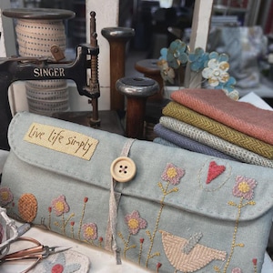 May include: A light blue fabric needle case with embroidered flowers, a bird, and the text "live life simply". The case has a wooden button and a white ribbon.