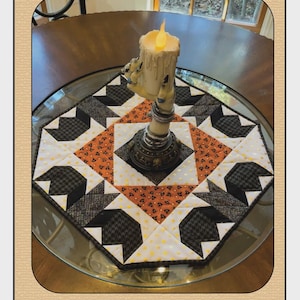 May include: A Halloween-themed quilted table runner with a central starburst design in orange, black, and white fabrics. The runner is topped with a candle holder shaped like a skeletal hand holding a candle. The text "Batusi" and "Quilted Garden Designs" are visible.
