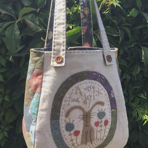 May include: A beige tote bag with a patchwork design. The front of the bag features a brown rabbit with the text "A very fine day" in a circle.