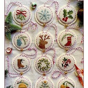 May include: Hand-embroidered Christmas ornament set featuring 12 unique designs. Each ornament is round with a scalloped edge and a small gold bell. Designs include a snowflake, holly, a reindeer, and a Christmas tree. The text "Deck the Boughs" is at the top.
