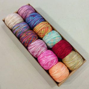 May include: A rectangular cardboard box filled with twelve colorful balls of yarn. The yarn colors include pink, blue, red, yellow, and green. The yarn is tightly wound into small, round balls. The box is sitting on a white surface.