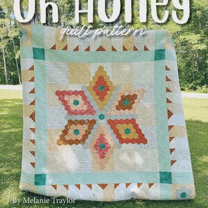 May include: A quilt featuring the Oh Honey quilt pattern by Melanie Traylor, a beginner English paper piecing project measuring 80 inches by 80 inches. The design displays a central star motif in orange, yellow, and teal, surrounded by script-printed fabric and a geometric border with small brown triangles.