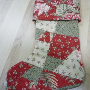 Op de afbeelding: Een kerstkous met een patchworkontwerp van rode en cr&egrave;mekleurige stof met bloemen- en bladpatronen. De boord is gemaakt van rode stof met een dichte bloemenprint en aan de bovenhoek zit een klein stoffen lusje om hem op te hangen.