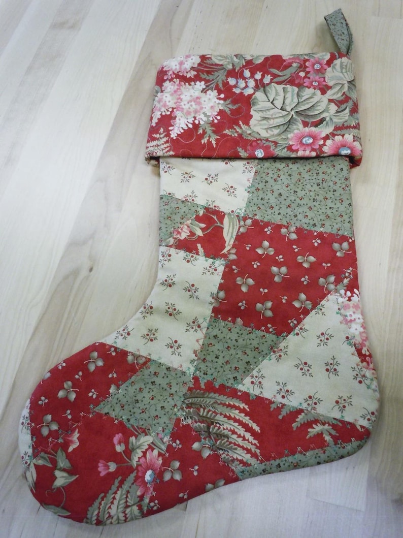 Op de afbeelding: Een kerstkous met een patchworkontwerp van rode en cr&egrave;mekleurige stof met bloemen- en bladpatronen. De boord is gemaakt van rode stof met een dichte bloemenprint en aan de bovenhoek zit een klein stoffen lusje om hem op te hangen.