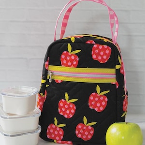 May include: A black quilted lunch bag featuring a red apple pattern with yellow leaves, a pink and white checkered handle, and a yellow zippered front pocket. The text "Lunch Box Delight" appears at the top, and a "KNOT + THREAD design" logo is at the bottom.