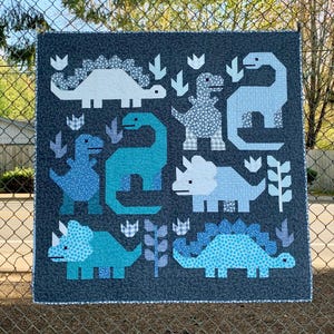 May include: A square quilt featuring a dark navy background with a white star pattern. The quilt displays various dinosaur shapes in shades of light blue, medium blue, and teal, accented with small white floral and checkered fabric patterns.
