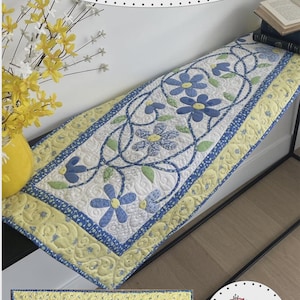 May include: A quilted table runner with a floral design. The runner features blue daisy-like flowers with green leaves on a white background, framed by a yellow and blue border. The finished size is 20" x 44". The text "Tangled Daisies" is at the top.