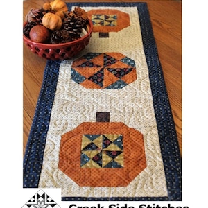 May include: A quilted table runner with a pumpkin patch design. The runner is 15" x 41" and features three orange pumpkin shapes with geometric patterns. The border is navy blue with white polka dots. The text "Pumpkin Patch" is at the top.