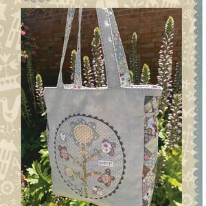 May include: A gray tote bag with floral and bird appliqués. The bag features a large, circular design with a tree and the words "love life." The handles and trim have a floral pattern. The text "Signs of Spring Bag" is at the bottom.