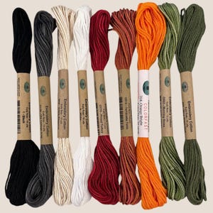 May include: A collection of embroidery cotton thread in various colors, including black, gray, white, red, orange, and green. Each skein is labeled with its color name and brand information. The threads are tightly wound and ready for use in needlework projects.