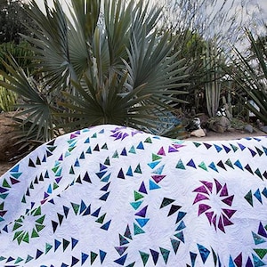 May include: White quilt with a repeating pattern of colorful triangles in shades of blue, green, and purple. The quilt has a spiral design and is set against a backdrop of desert plants and a cloudy sky. The text "Quiltworx.com" is in the upper right corner.