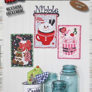 May include: A display featuring winter-themed fabric art, including a snowman in a mug, a gingerbread man, and a cupcake. The text "WINTER NIBBLERS" is displayed above the art pieces. The finished size of each piece is 6" x 9".