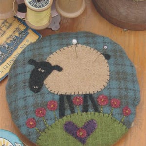 May include: A round, blue and tan sheep pin cushion with a felt sheep design. The sheep has a tan body, black head and legs, and is surrounded by red flowers and a purple heart. A pearl-headed pin is inserted.