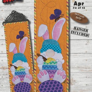 May include: Appliqued fabric wall hanging featuring a gnome with bunny ears, holding a paintbrush and an Easter egg. The design is 6" x 20" and includes the text "Apr #4 of 12". The skill level is easy, and a hanger is included.