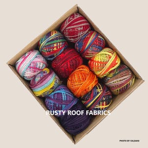 May include: A cardboard box filled with colorful yarn balls. The yarn balls are various colors, including red, orange, yellow, blue, and purple. The text "RUSTY ROOF FABRICS" is visible in the image.