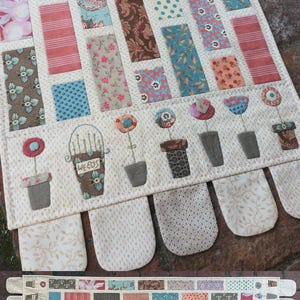 May include: A patchwork quilt runner with a brick pattern in various shades of brown, pink, blue, and green. The runner has a border of white fabric with a floral pattern. The bottom edge of the runner has a row of fabric flowers in pots.