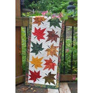 May include: A quilted table runner featuring a pattern of colorful autumn leaves in shades of red, green, yellow, and brown. The leaves are arranged on a white background with a quilted pattern, bordered by a patterned fabric.