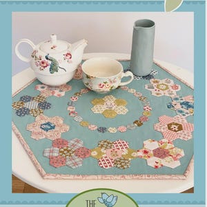May include: A floral teapot, teacup, and pitcher sit on a round table with a blue floral patterned table topper. The table topper features a floral design with pink, blue, and yellow fabric. The text "Teatime Tabletopper" is at the top.
