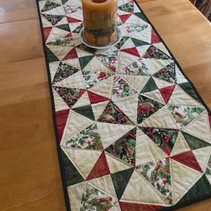 May include: A quilted table runner with a geometric design featuring red, green, and cream-colored triangles. The runner has a black border and is decorated with a festive bird and berry pattern. A large, cylindrical candle sits on top.