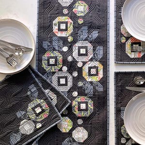 May include: A black quilted table runner and four matching placemats. The set features a floral design with geometric shapes in shades of gray, green, and pink. The text "POSIES Table Runner and 4 Placemats" is displayed at the top.