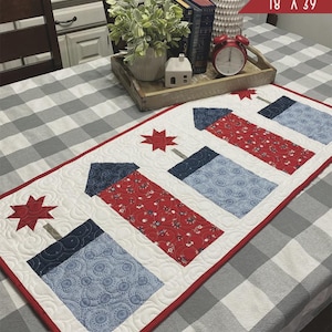 May include: A quilted table runner with a firework design. The runner is white with red and blue firework shapes and star accents. The text "FIREWORK FRENZY" and "PRIMROSE COTTAGE" are displayed. The runner measures 18" x 39".