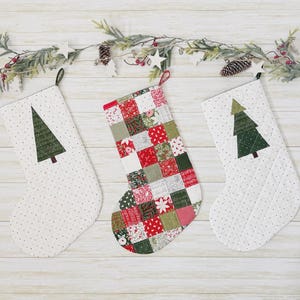 May include: Three Christmas stockings hanging in a row. The two outer stockings are white with small green dots and feature a single green fabric Christmas tree applique. The center stocking has a patchwork design of red, green, and white fabric squares.