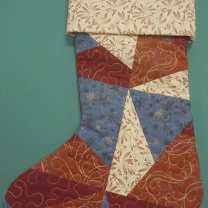 Op de afbeelding: Een gewatteerde kerstkous met een patchworkontwerp van driehoekige stofsegmenten in tinten donkerrood, gedempt blauw en cr&egrave;me met een botanische print. De boord is cr&egrave;me met een blad- en bessenpatroon en een donkerrode lus om op te hangen.