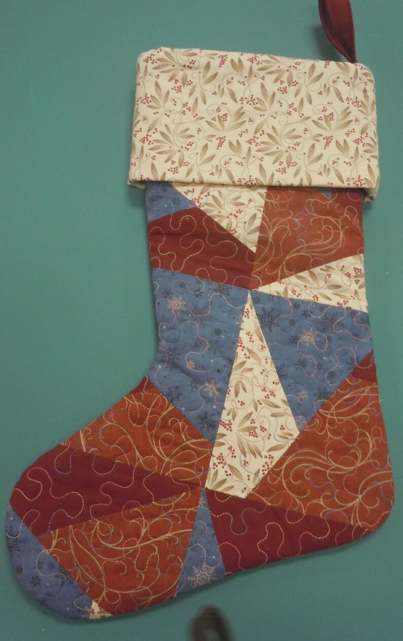 Op de afbeelding: Een gewatteerde kerstkous met een patchworkontwerp van driehoekige stofsegmenten in tinten donkerrood, gedempt blauw en cr&egrave;me met een botanische print. De boord is cr&egrave;me met een blad- en bessenpatroon en een donkerrode lus om op te hangen.