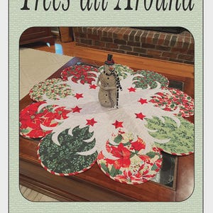 May include: A round, quilted table topper with a Christmas tree and poinsettia design. The topper is white with red and green accents, and features a snowman figurine in the center. Text on the image reads "Trees all Around" and "Quilted Garden Designs".