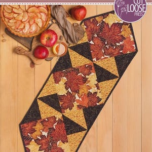 May include: A Jubilee Runner quilt pattern designed by Cathey Laird, featuring a table runner with a maple leaf print in red, orange, and yellow, accented by black and gold geometric triangles. The pattern measures 12 inches by 36 inches.
