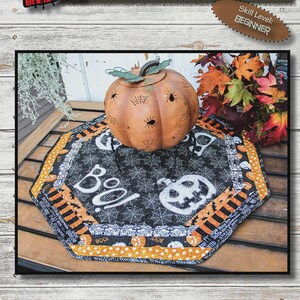 May include: Halloween-themed table mat, 22-inch octagon, with the text "BOO YA! TABLE MAT". The mat features a black background with white spiderwebs and a jack-o'-lantern design. A decorative pumpkin sits on top. Skill level: Beginner.