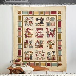 May include: A beige quilt with the word "SEW" in the center, surrounded by sewing-themed images. The quilt measures 56" x 68". The top left corner has the "BUTTERMILK BASIN DESIGN CO." logo. The quilt is displayed on a wall.