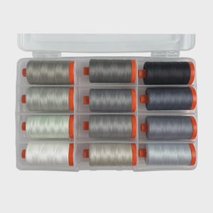 May include: A clear plastic organizer filled with spools of thread in various shades of gray, black, and white. Each spool has orange end caps. The thread is neatly arranged in rows within the clear container, suggesting a sewing or crafting theme.