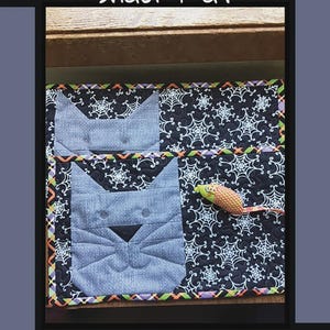 May include: A rectangular quilted snack mat featuring a black fabric background with a white spiderweb pattern. The mat includes two light blue cat face appliques and a small fabric mouse toy. The border has a colorful orange, purple, and green pattern.