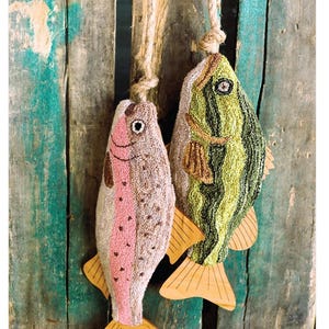 May include: Two colorful fish-shaped needlepoint ornaments, a green bass and a pink trout, hang from a rope on a weathered wooden background. The fish have wooden tails and are labeled "Fish Tails Bass & Trout".