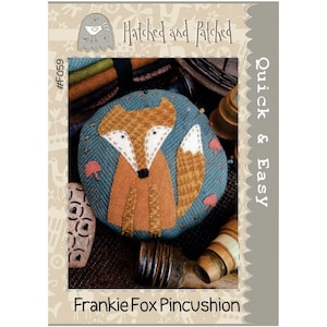 May include: A brown and orange felt fox pincushion with a blue felt background. The pincushion is round and has a small mushroom design on the background. The pincushion is labeled "Frankie Fox Pincushion" and "Quick & Easy" with the number "#F059".