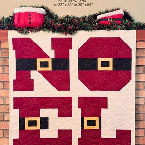 May include: A Christmas-themed quilt pattern with the text "PÈRE NOËL" in green. The quilt design spells out "NOËL" in red with black belts and gold buckles. The pattern finishes at 42" x 42" or other sizes. The text "MY BESTE QUILT CO" is also visible.
