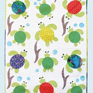 May include: A white quilt featuring a pattern of sea turtles in various colors and patterns. The turtles have green flippers and heads, with shells in red, green, yellow, and blue floral designs. The quilt includes the text "Sea Turtle Friends" and "Applique Quilt Sewing Pattern".